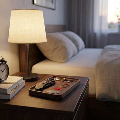 Wooden nightstand with lamp, books, and tray with keys and coins in a bedroom setting.